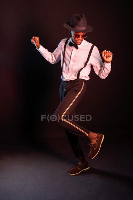 Young masculine ethnic male model in hat and trousers standing dancing while looking away on black background — Stock Photo