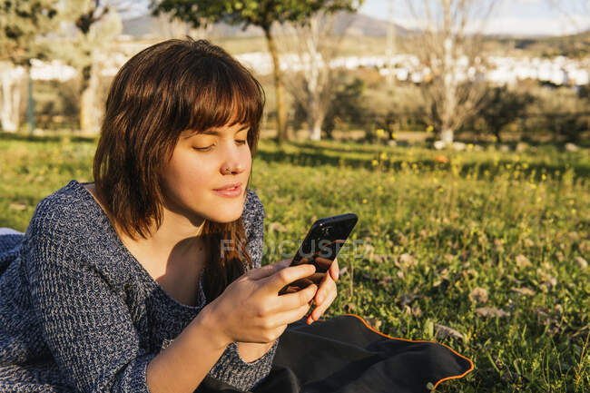 Mujer despreocupada acostada sobre cuadros en el prado y usando un teléfono inteligente mientras disfruta de un picnic en un día soleado en primavera - foto de stock