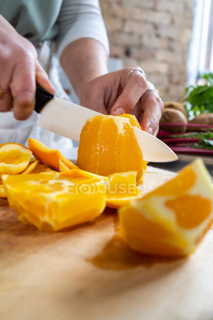 Cultivo irreconocible corte femenino naranjas jugosas maduras con cuchillo en tabla de cortar de madera en la mesa de la cocina - foto de stock
