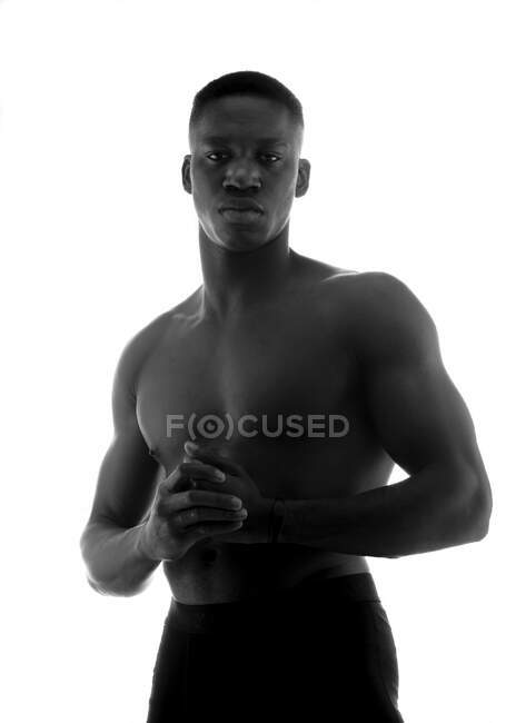 Schwarz-Weiß eines emotionslosen, muskulösen jungen schwarzen Mannes mit gefalteten Händen, der im Studio auf weißem Hintergrund in die Kamera blickt — Stockfoto