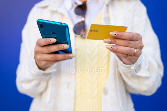 Cultivado irreconocible mujer afroamericana elegante que paga con tarjeta de plástico durante las compras en línea a través del teléfono móvil, mientras que de pie sobre fondo azul en el estudio - foto de stock