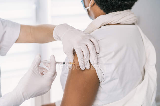 Verkorkste Ärztin in Schutzuniform, Latexhandschuhen und Gesichtsmaske, die anonyme afroamerikanische Patientin in der Klinik während des Coronavirus-Ausbruchs impft — Stockfoto