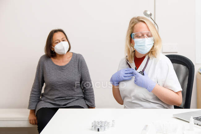 Especialista médica femenina en uniforme protector y guantes de látex vacunando a una paciente mayor en la clínica durante el brote de coronavirus - foto de stock