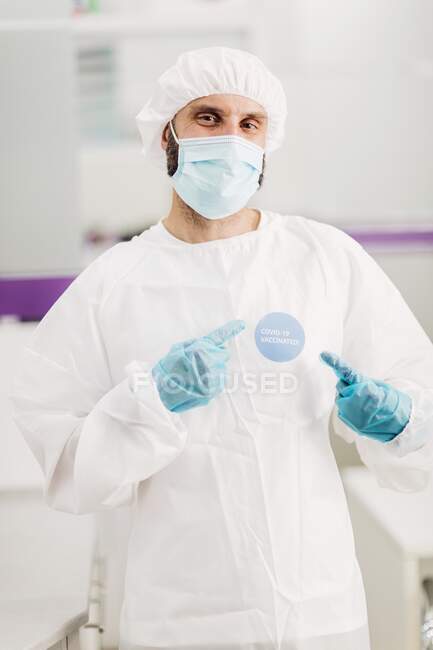 Позитивный врач-мужчина в латексных перчатках и защитной медицинской маске указывает на привитую наклейку ковид-19 на белой униформе, стоящей в современном медицинском кабинете и смотрящей в камеру — стоковое фото