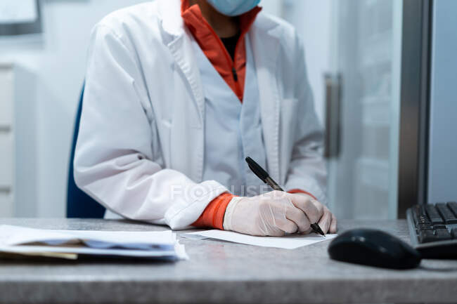Неузнаваемая женщина-врач в маске и форме сидит за столом в медицинской комнате и выписывает рецепт на бумаге — стоковое фото
