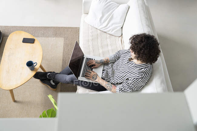 De cima jovem hipster macho com cabelos cacheados navegando na internet no netbook enquanto descansa no sofá na sala da casa — Fotografia de Stock
