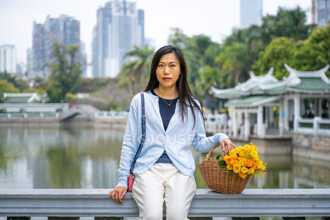 Menina da bela asiática retrato em um parque, enquanto ela se senta ao lado de cesta de vime com flores amarelas. — Fotografia de Stock