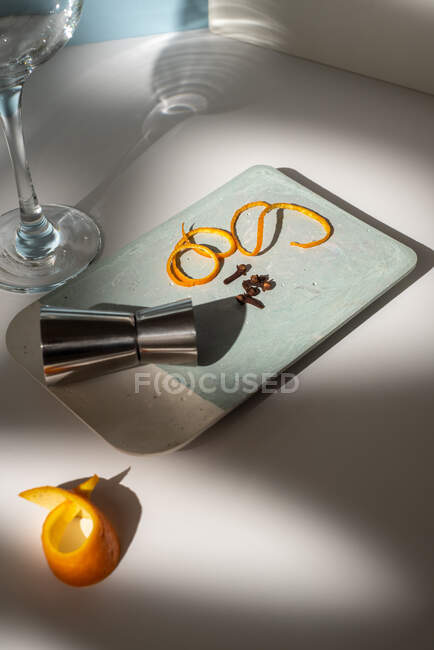 From above of glass near metal jigger with citrus fruit zest and dry condiments on table with shadows — Stock Photo