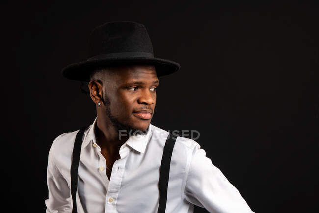 Young masculine ethnic male model in hat and trousers standing while looking away on black background with smoke — Stock Photo
