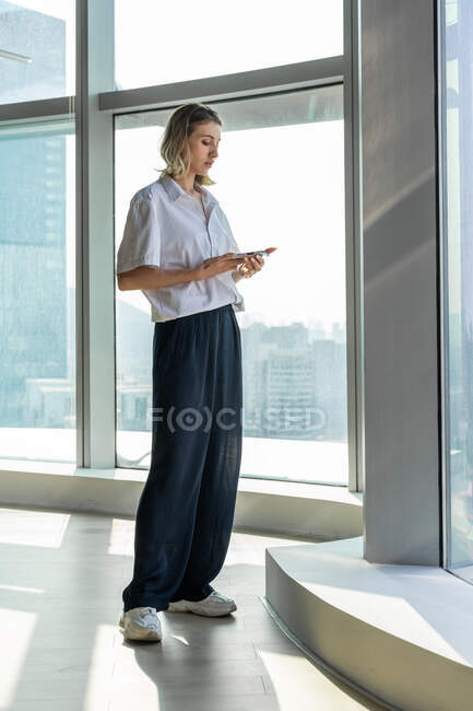 Jovem solitária e pouco emocional de pé no escritório vazio com grande janela navegando no telefone celular — Fotografia de Stock