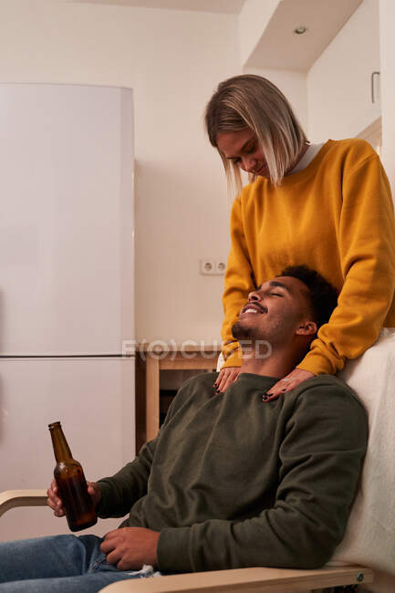 Vista lateral namorada dando namorado massagem enquanto ele está desfrutando de uma cerveja sentada em poltrona em casa — Fotografia de Stock