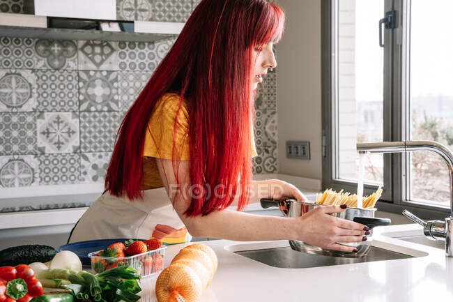 Jeune homosexuelle femelle versant de l'eau du robinet dans une casserole avec des pâtes non cuites contre divers légumes dans la maison — Photo de stock