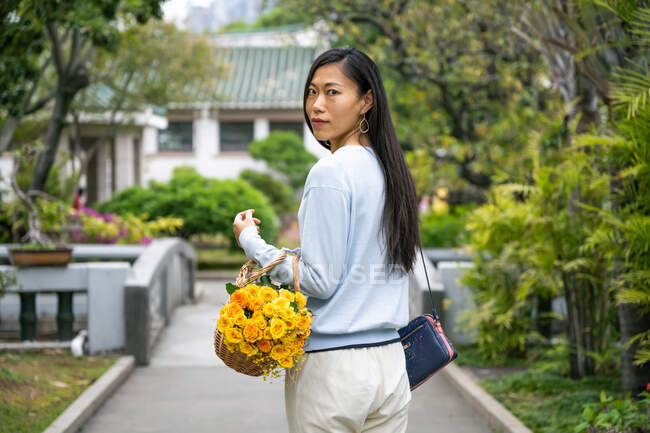 Das Mädchenporträt der schönen Asiatin in einem Park, während sie einen Weidenkorb mit gelben Blumen trägt. — Stockfoto