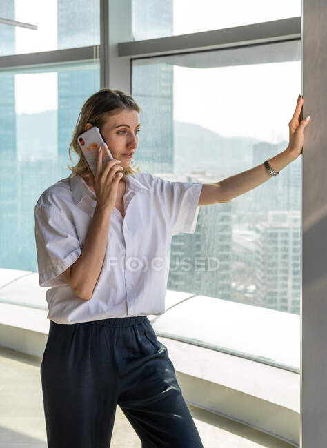 Junge glückliche Geschäftsfrau steht im Büro mit großen Fenstern und telefoniert mit dem Handy — Stockfoto