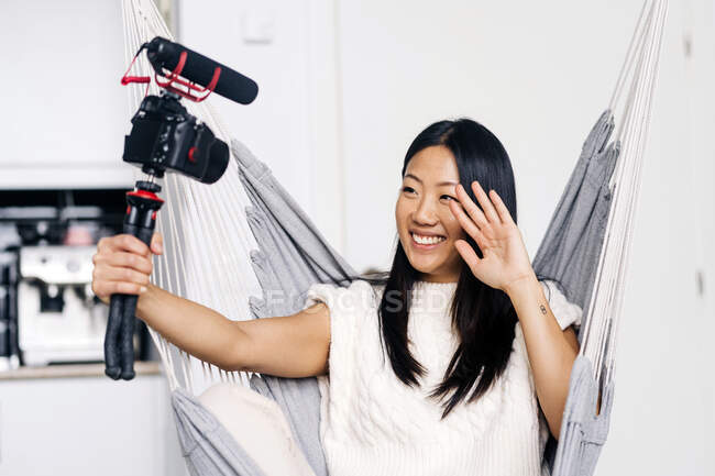 Smiling ethnic female vlogger recording video on photo camera while sitting on hammock in the living room — Stock Photo