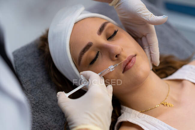 Сверху неузнаваемый профессиональный косметолог со шприцем, инъекционным наполнителем с гиалуроновой кислотой в губы клиентки во время процедуры в клинике красоты — стоковое фото