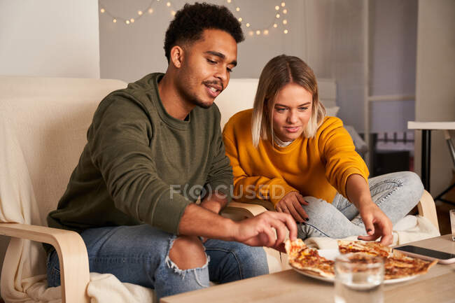 Multiethnisches Paar sitzt in Sesseln und isst leckere Pizza, während es das Wochenende zu Hause zusammen genießt — Stockfoto