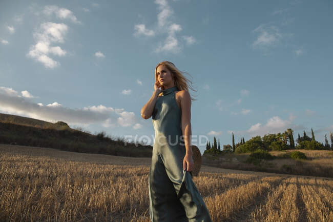 Mulher pacífica em vestido elegante em pé no campo seco na área rural e olhando para longe — Fotografia de Stock