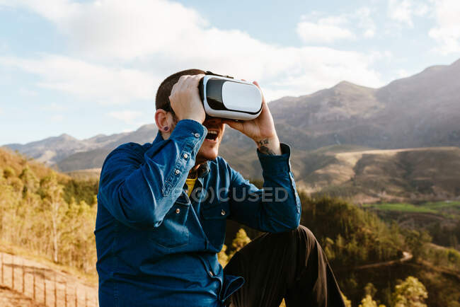 Curious male traveler sitting on hill and experiencing virtual reality in goggles in mountains on sunny day — Stock Photo