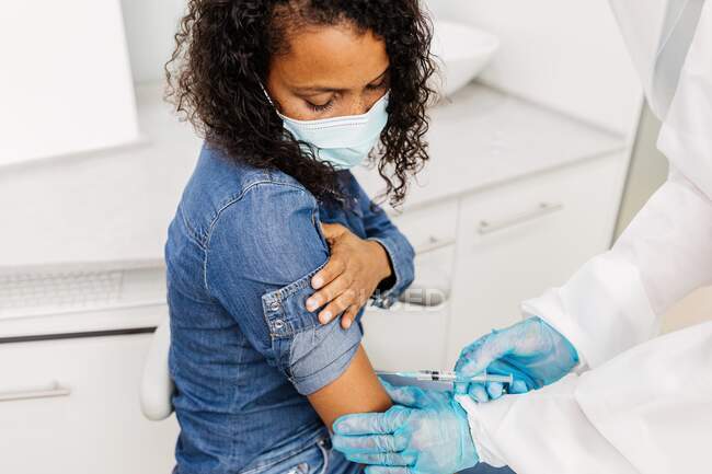 Specialista medico maschile irriconoscibile ritagliato in uniforme protettiva e guanti di lattice che vaccinano la paziente afroamericana in clinica durante l'epidemia di coronavirus — Foto stock