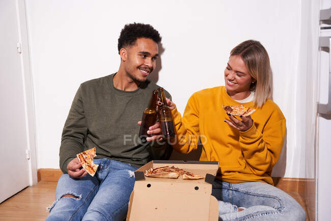 Alegre pareja multiétnica sentada en el suelo mientras comen deliciosa pizza y beben cerveza juntos - foto de stock