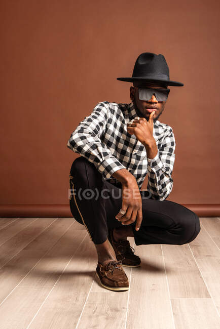 Cooles afroamerikanisches Model in kariertem Hemd und Hut, das in die Kamera schaut, während es auf braunem Hintergrund hockt — Stockfoto