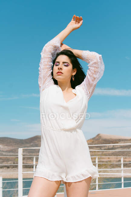 Молода ніжна брюнетка жінка з макіяжем і закритими очима в білому, стоячи в паркані проти річки під блакитним хмарним небом — стокове фото