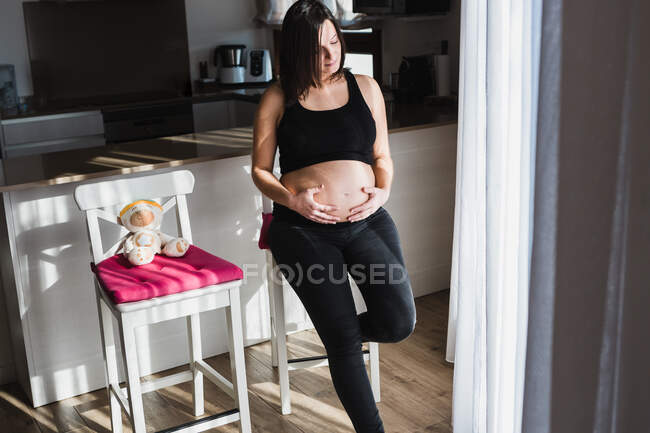 Высокий угол безмятежной беременной женщины, нежно касающейся живота, стоя возле стула дома — стоковое фото