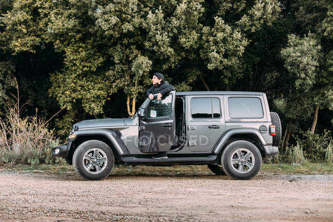 Side view of an adventurer leaning on the door of an off-road car while looking away before starting the journey — Stock Photo