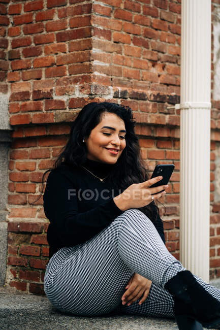 Deleitado más tamaño femenino en traje de moda sentado en la frontera de piedra en la ciudad y mensajería en el teléfono móvil - foto de stock