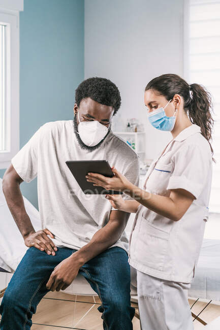 Doctora joven en uniforme médico y estetoscopio con mascarilla facial que habla y muestra el resultado en la tableta al paciente afroamericano durante la cita en la clínica - foto de stock