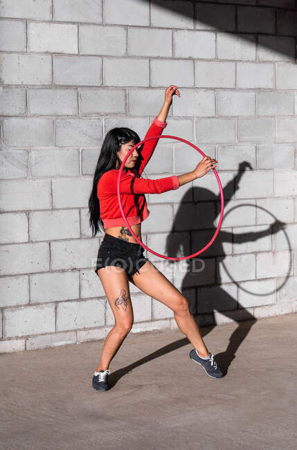 Joven mujer tatuada en ropa deportiva girando hula hoop mientras baila contra paredes de ladrillo con sombras y mirando hacia adelante a la luz del sol - foto de stock