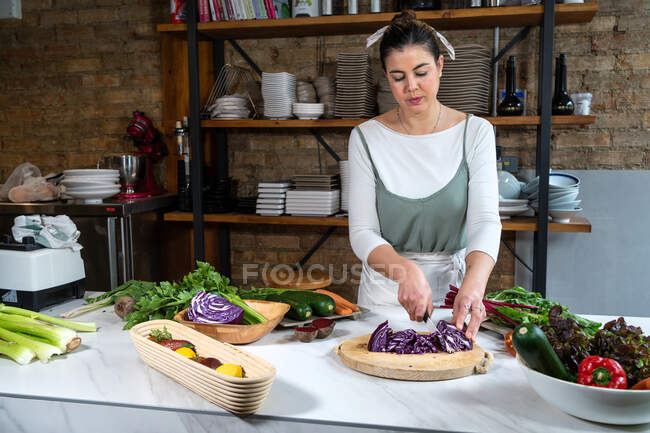 Взрослая женщина режет красную капусту ножом во время приготовления вегетарианской пищи за столом в лофт-хаусе — стоковое фото