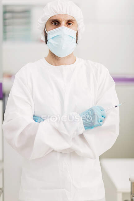 Позитивний лікар чоловічої статі з латексними рукавичками та захисною медичною маскою з я щеплюю повідомлення наклейка на білій уніформі, що стоїть з обіймами, схрещеними в сучасному медичному кабінеті та дивлячись на камеру — стокове фото