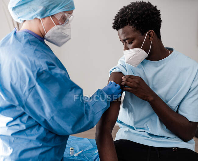 Vista laterale del medico donna in uniforme protettiva e guanti di lattice che disinfettano il braccio con cotone con alcol per vaccinare il paziente afroamericano in clinica durante l'epidemia di coronavirus — Foto stock