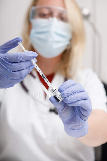 Розмиті невизначені ліки в захисній масці для обличчя та латексні рукавички з флаконом коронавірусної вакцини та шприцом, що показують камеру, стоячи в лікарняній кімнаті — стокове фото