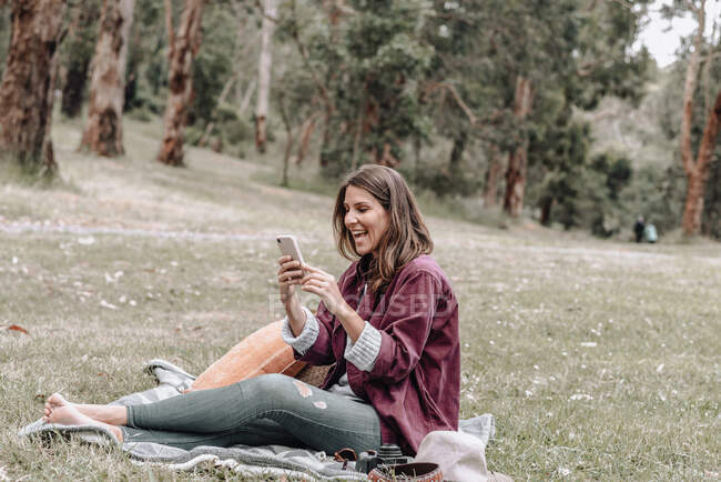 Cheerful female in hat sitting on blanket on meadow in forest and browsing mobile phone while enjoying picnic in Australia — Stock Photo