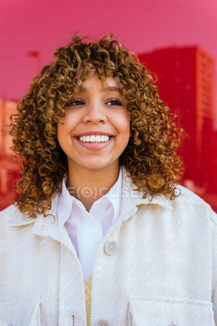 Mulher afro-americana encantada com penteado encaracolado em pé no fundo vermelho no estúdio olhando para longe — Fotografia de Stock