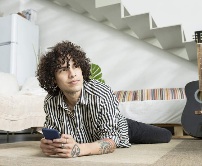 Junger kontemplativer Mann im gestreiften Hemd mit Tätowierungen und Handy auf Teppich liegend, während er in der Wohnung nach oben schaut — Stockfoto