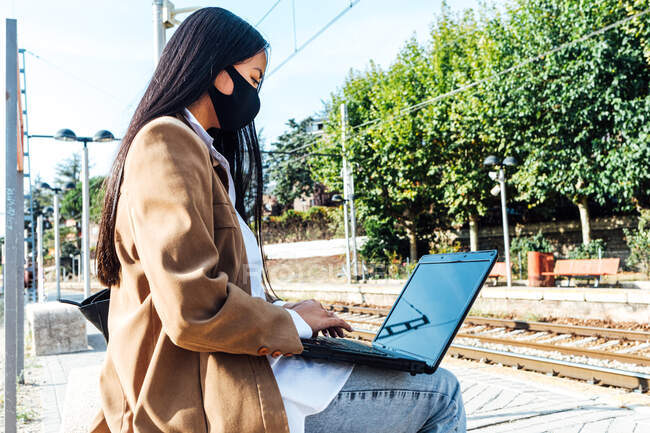 Vista laterale della femmina asiatica in maschera seduta sulla panchina della stazione ferroviaria e portatile di navigazione in attesa del treno — Foto stock