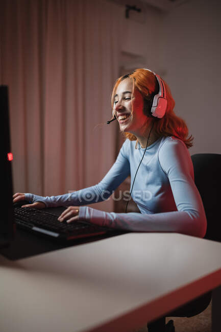 Vista lateral del jugador femenino encantado en auriculares jugando videojuego mientras está sentado en la mesa en casa - foto de stock