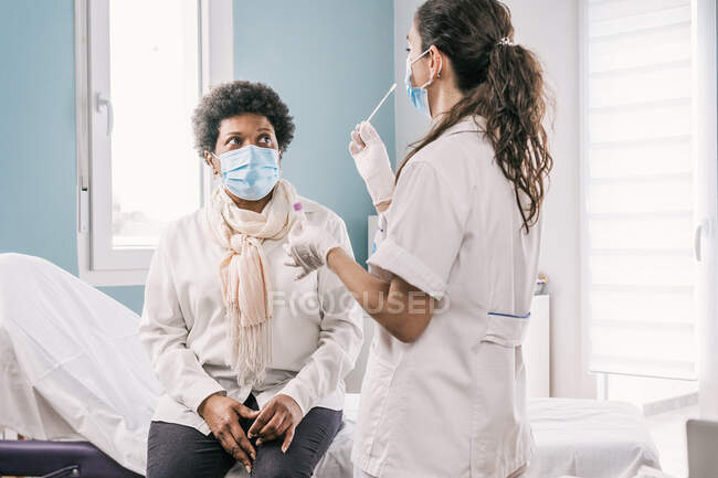 Female medical specialist in protective uniform, latex gloves and face mask doing nasal coronavirus test on African American mature woman patient in clinic during virus outbreak — Stock Photo