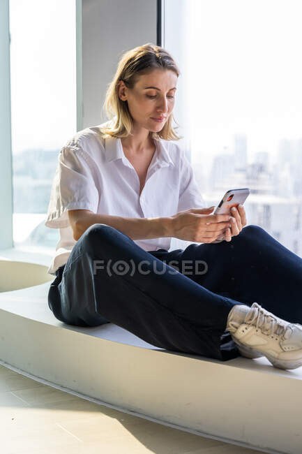 Solitario unemotional giovane donna seduta in ufficio vuoto con grande finestra di navigazione sul telefono cellulare — Foto stock