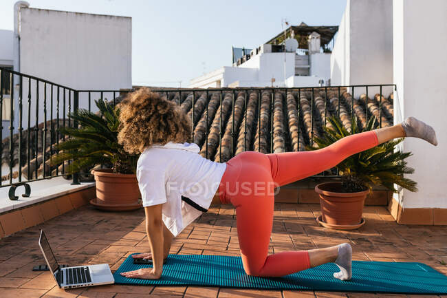 Vista lateral de ajuste femenino negro equilibrio en pose de mesa mientras se practica yoga en la azotea y ver lección en línea en el ordenador portátil - foto de stock