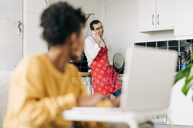 Hombre en delantal hablando en el teléfono inteligente en la cocina y la mujer negra portátil de navegación en casa - foto de stock