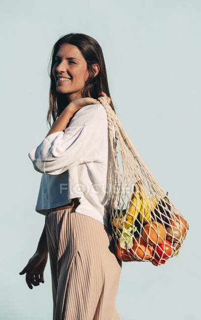 Side view of smiling female standing with assorted fruits and vegetables in eco friendly mesh bag against blue wall in city and looking away — Stock Photo
