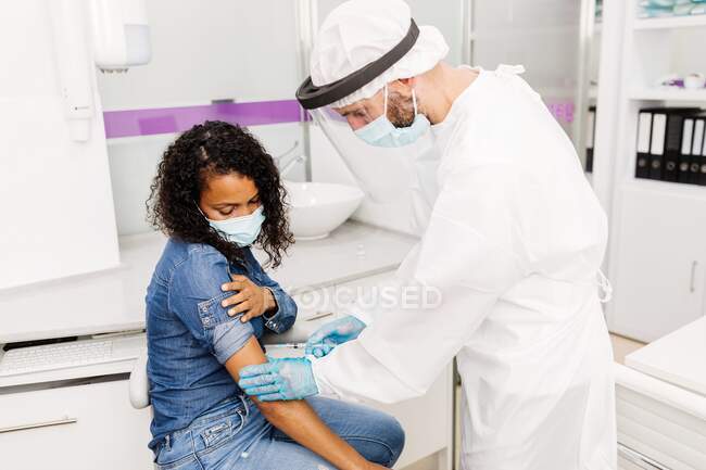 Vista lateral del especialista médico masculino en uniforme protector, guantes de látex y protector facial vacunando a la paciente afroamericana en la clínica durante el brote de coronavirus - foto de stock