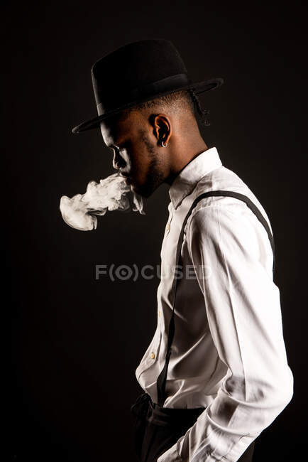 Бічний погляд на чоловіка афроамериканського походження в білій сорочці і капелюсі, який видихає пару, курюючи сигарету. — стокове фото