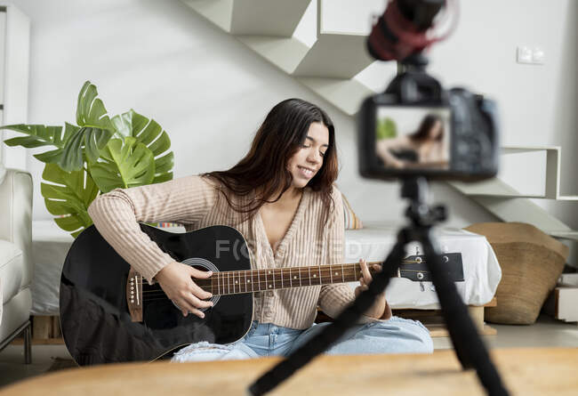 Молода жінка-гітаристка грає на акустичній гітарі під час запису відео на фотокамеру в домашній кімнаті — стокове фото