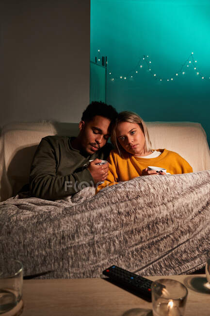 Многорасовая пара, сидящая под одеялом в креслах с чашками горячих напитков и глядя в сторону во время отдыха в уютной гостиной — стоковое фото
